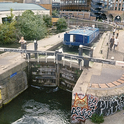 The image features a river with a bridge crossing over it. On the other side of the river, there is a city street lined with buildings and people walking around. A few individuals are also seen near the water's edge, possibly enjoying the view or taking a stroll.