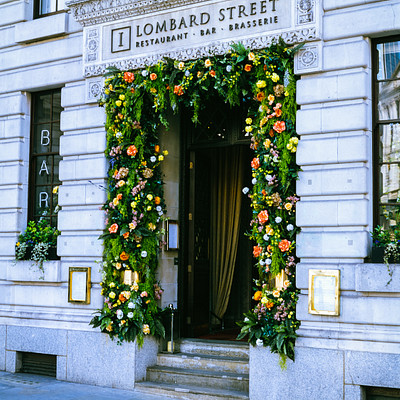 The image features a beautifully decorated entrance to the Lombar Street Bar. The doorway is adorned with two large flower garlands, adding an elegant touch to the building's facade. Above the door, there are several potted plants that further enhance the overall appearance of the establishment.