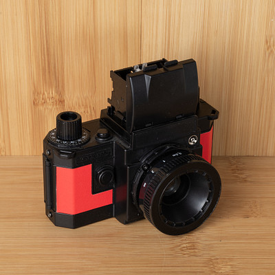 The image features a vintage camera sitting on top of a wooden table. The camera is black and red in color, with the lens facing upwards. It appears to be an old-fashioned model, possibly a Polaroid or a similar type of camera.