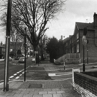 The image is a black and white photo of an empty street in the city. There are several cars parked along the side of the road, with some closer to the foreground and others further away. A few people can be seen walking on the sidewalk or standing near the edge of the street.