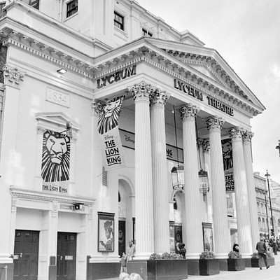 The image is a black and white photo of the Lyceum Theatre, which features a large white building with columns. There are several people walking around outside the theatre, some closer to the foreground while others are further away. A few individuals can be seen standing near the entrance of the theatre.