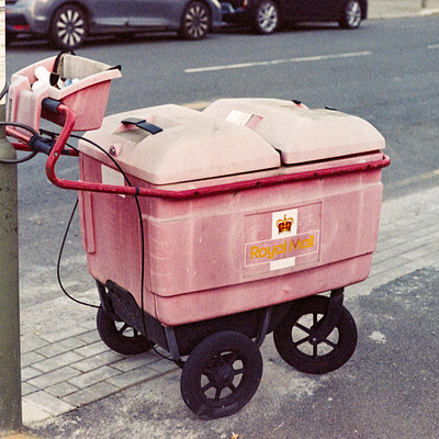 The image features a pink trash can with an attached cart, parked on the side of a street. There are two cars visible in the scene; one is located behind the trash can and another car is further away to the right. A parking meter is also present near the left edge of the image.
