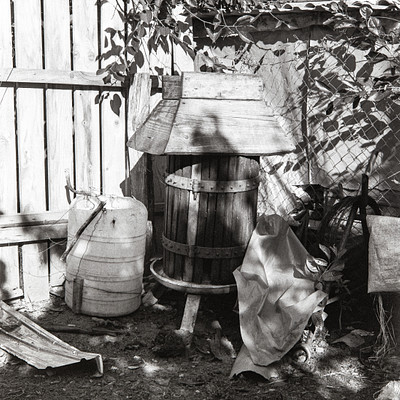 The image is a black and white photo of an outdoor scene with various items scattered around. There are two barrels, one large and one small, placed next to each other in the center of the scene. A wooden bucket can be seen nearby, along with a potted plant on the right side of the frame.