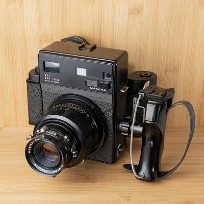 The image features an old-fashioned camera sitting on a wooden table. It is placed next to a cell phone, creating a contrast between the two devices. The camera has a strap attached to it, and its lens is visible in the foreground of the photo.