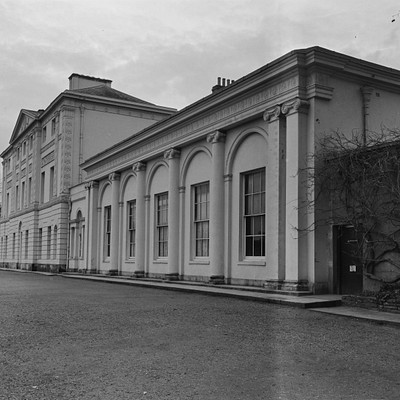 The image is a black and white photo of an old building, possibly a mansion or a large house. It has several windows on the front side, with some located near the top floor and others closer to the ground level. A doorway can be seen in the middle of the building, leading inside.