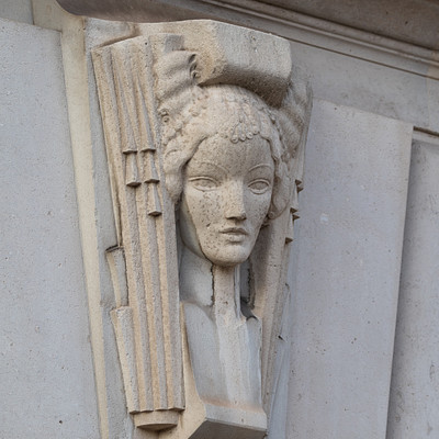 The image features a white building with an intricate carved stone design on the side. This artistic piece showcases a woman's face, possibly depicting a goddess or a mythical figure. The carving is set into the wall and appears to be part of a larger architectural structure.
