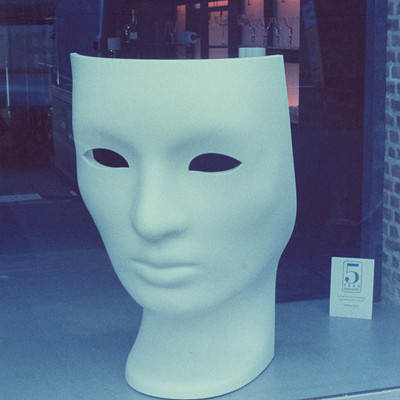 The image features a large white mask on display in front of a window. The mask is positioned prominently, covering most of the frame. In addition to the mask, there are several books scattered around the area, with some placed near the top left corner and others closer to the center of the scene.