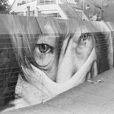 The image is a black and white photo of a mural on the side of a building. The mural features a woman with her eyes covered, possibly by a mask or blindfold. She appears to be looking over her shoulder at something in the distance.