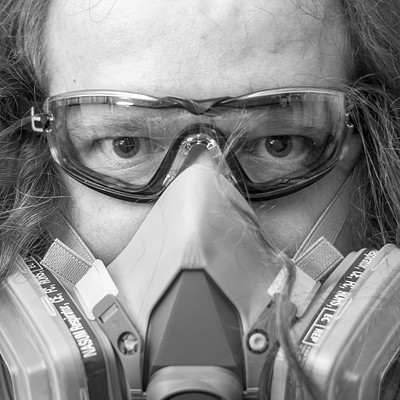 The image features a man wearing safety goggles and a gas mask, possibly for protection against hazardous materials. He is looking directly at the camera with an intense expression on his face. His eyes are visible through the protective glasses, adding to the overall intensity of the scene.