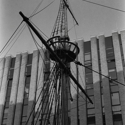 The image is a black and white photo of an old sailing ship, possibly a pirate ship, docked in front of a building. The ship has a large mast with sails attached to it, giving the impression that it's ready for a journey or has just returned from one. The building behind the ship is made of brick and features multiple windows on its facade.