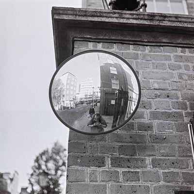 The image is a black and white photo of a mirror mounted on the side of a brick building. The reflection in the mirror shows a street scene with several people walking by, including one person sitting down. There are also two cars visible in the reflection, one parked further back and another closer to the foreground.