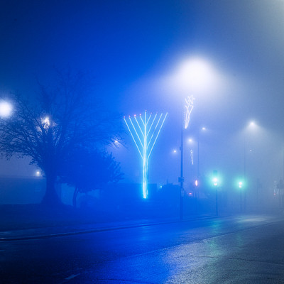 The image depicts a foggy night scene with a blue hue, creating an atmospheric ambiance. There are several street lights illuminating the area, casting a soft glow on the surroundings. A large tree is visible in the foreground, adding to the overall mood of the scene.