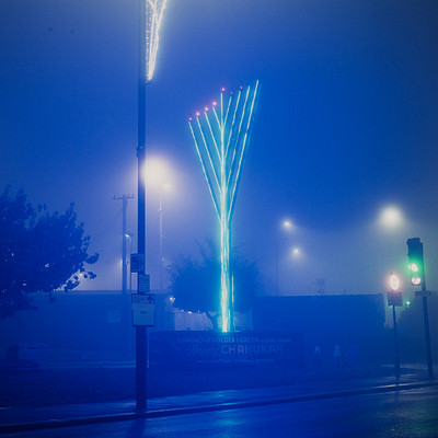 The image is a nighttime city scene with a blue sky and foggy atmosphere. There are several traffic lights visible in the area, including one on the left side of the street, another further down the road, and two more towards the right side. A large blue light pole stands out in the middle of the scene, adding to the overall ambiance.
