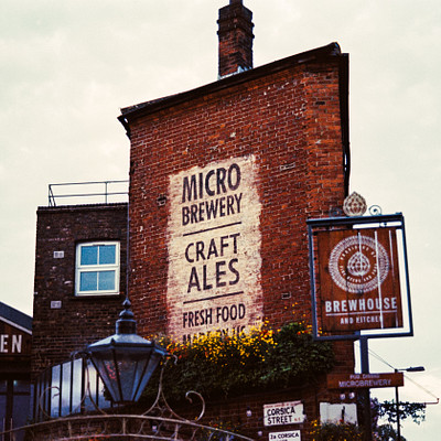 The image features a brick building with a large sign on its side, advertising "Micro Brewery." Above the brewery sign is an old-fashioned lamp post. In front of the building, there are two potted plants adding greenery to the scene. Additionally, there are several people in the image, with one person standing near the left side and another person closer to the right side of the frame.