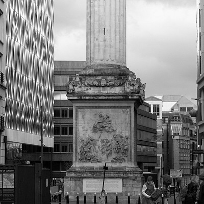 The image is a black and white photo of a city street with an old monument in the center. A tall tower, possibly a clock tower or a monument, stands prominently on the street corner. There are several people walking around the area, some closer to the monument while others are further away.