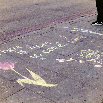 The image is a black and white photo of a sidewalk with various writings on it. There are several chalk drawings, including one that says "There is more to care." Another drawing features a heart and the words "I love you." A person can be seen standing near the sidewalk, possibly admiring or participating in creating these artistic expressions. The scene captures a moment of creativity and connection with others through public art.