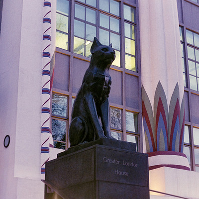 The image features a statue of a cat sitting on top of a pedestal, located in front of a building. The cat statue is positioned near the center of the scene and appears to be the main focus of the image. The building itself has multiple windows, with some situated towards the left side of the image and others on the right side.