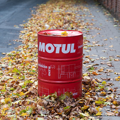 The image features a red and white Motul barrel sitting on the side of a road. It is surrounded by fallen leaves, creating an autumn atmosphere. The barrel appears to be empty, with no trash inside it.