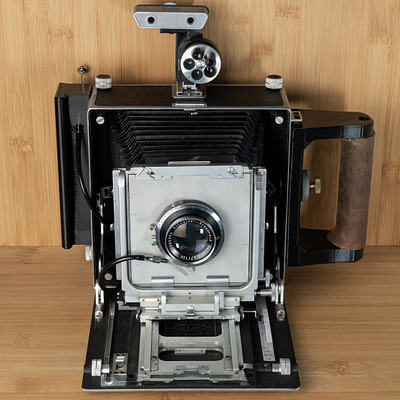 The image features an old-fashioned camera sitting on a wooden table. It is an antique model with a black and silver color scheme, giving it a vintage appearance. The camera has a lens in the front, which is visible as part of its design.