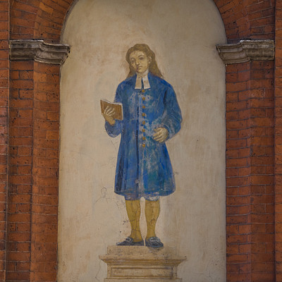 The image features a man dressed in blue clothing, standing on a pedestal and holding a book. He appears to be an important figure or possibly a religious leader, as he is wearing a cape and a collar. The scene takes place against the backdrop of a brick wall, which adds texture and depth to the image.