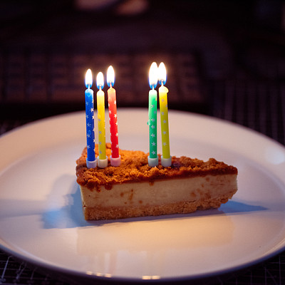 The image features a white plate with a slice of cake on it. The cake is decorated with candles, and there are five candles in total, each one placed at different positions around the cake. The arrangement creates an appealing presentation for the dessert.