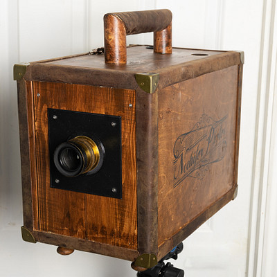 The image features a wooden box with a metal handle, which appears to be an old-fashioned camera. It is mounted on the wall and has a lens cover attached to it. The box seems to be made of wood and leather, giving it a vintage appearance.