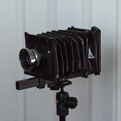 The image features a black camera with a lens on it, placed on top of a tripod. The camera is positioned in front of a white wall, and the tripod legs are visible beneath it. The scene appears to be set indoors, possibly for photography or videography purposes.