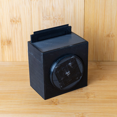 The image features a small black box sitting on top of a wooden table. The box is made from wood and has a hinged lid, which is currently closed. It appears to be an old-fashioned design with a unique appearance.
