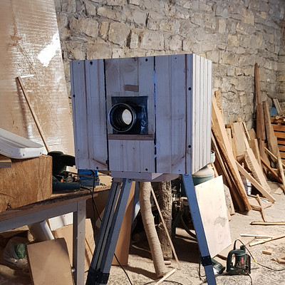 The image features a wooden box with a camera mounted on top of it. The camera is positioned in the center of the scene, and there are several other objects surrounding it. In addition to the camera, there are multiple tools scattered throughout the area, including scissors located near the left side of the frame and a pair of pliers closer to the right side.