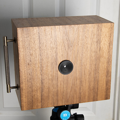 The image features a wooden box with a blue circle on it, which is mounted to the wall. It appears to be an old-fashioned camera or possibly a vintage photo holder. The box has a metal bracket attached to its back, allowing for easy mounting and display.