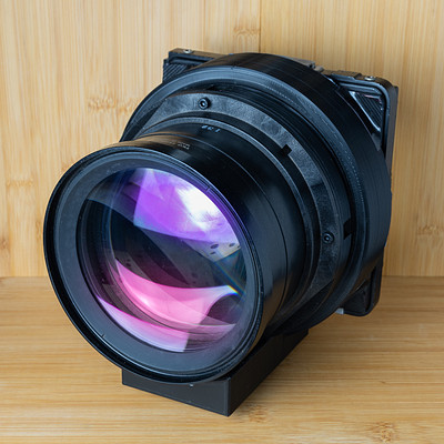 The image features a large, black camera lens with purple and pink tinted glass. It is placed on top of a wooden table or counter, possibly for display or as part of an artistic setup. The lens appears to be the focal point of the scene, drawing attention due to its unique coloring and size.