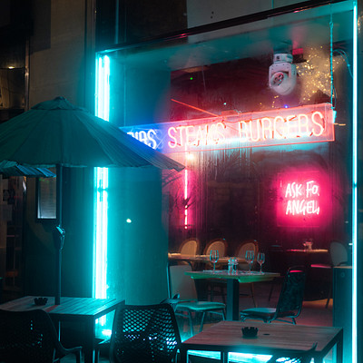 The image features a restaurant with an outdoor seating area. There are several tables and chairs arranged outside, some of which have umbrellas providing shade for the guests. A neon sign is visible above the dining area, adding to the ambiance of the place.