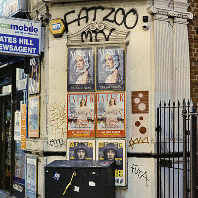 The image features a storefront with a black trash can in front of it. The trash can is covered in flyers and advertisements, making it an eye-catching display for passersby. There are several signs on the building as well, including one that says "Fat 20." Above the trash can, there are posters featuring a woman with a hat.