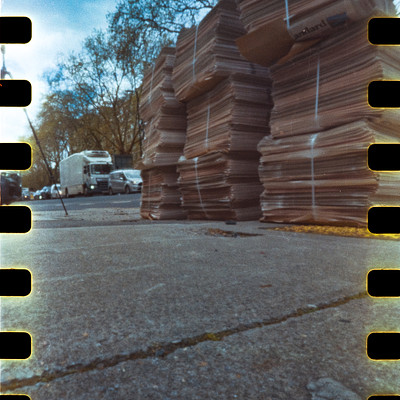 The image is a black and white photo of a pile of brown paper, possibly cardboard boxes or newspapers. There are several stacks of these items, with some placed on the ground and others sitting on top of each other. A truck can be seen in the background, parked near the pile of paper.