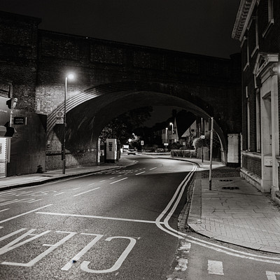 The image is a black and white photo of an empty street at night. There are several cars parked along the side of the road, with some closer to the foreground and others further away. A few people can be seen walking on the sidewalk or standing near the edge of the street.