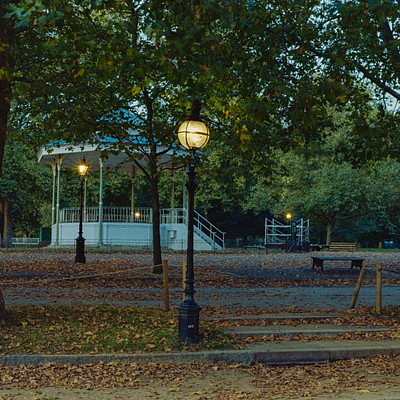 The image features a park with a gazebo and a fenced-in area. There are several benches placed around the park, providing seating for visitors to relax and enjoy the surroundings. A street light is present in the scene, illuminating the area at night.