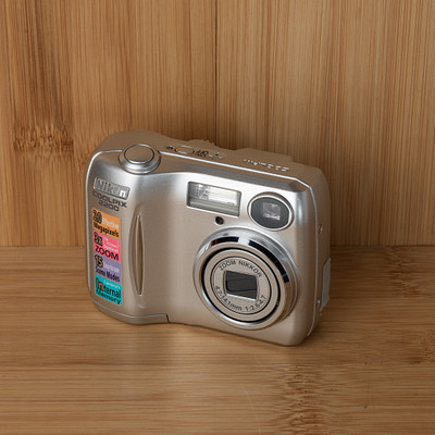 The image features a silver digital camera sitting on top of a wooden table. The camera is placed in the center of the scene, and it appears to be an older model with a few buttons visible on its surface. The wooden table provides a natural backdrop for the camera, creating a sense of warmth and simplicity.