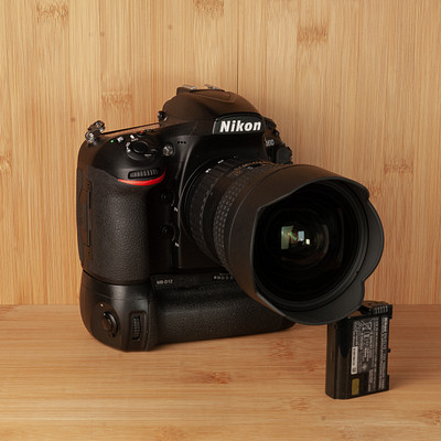 The image features a black Nikon camera with its lens open, sitting on top of a wooden table. The camera is positioned in the center of the frame and appears to be ready for use. The lens is large and occupies most of the lower portion of the image.