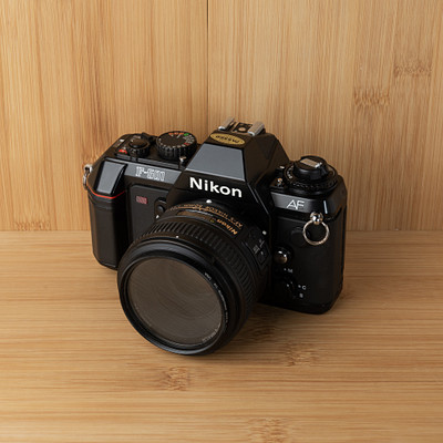 The image features a black Nikon camera sitting on top of a wooden table. The camera is placed in the center of the scene, with its lens facing upwards towards the viewer. The wooden surface provides an appealing backdrop for this classic piece of photography equipment.