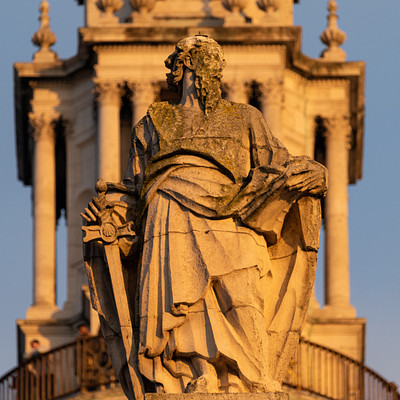 The image features a statue of a man holding two swords, standing on top of a building. The statue is positioned in the center of the scene and appears to be made of stone or concrete. The building has a clock tower with a bell at its base, adding an architectural element to the structure.
