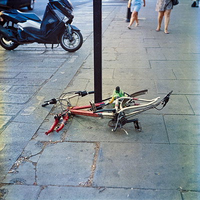 The image features a bicycle parked on the sidewalk next to a pole. There are two people walking by, one closer to the left side of the scene and another further back on the right side. A motorcycle is also visible in the background, positioned behind the bicycle.