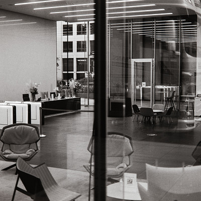The image is a black and white photo of an office building lobby. There are several chairs placed throughout the room, with some near the center and others closer to the edges. A dining table can be seen in the middle of the space, surrounded by chairs.