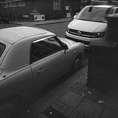 The image is a black and white photo of two cars parked on the side of a street. One car is a silver Volkswagen, while the other is a tan convertible. Both cars are parked next to each other, with the silver Volkswagen being closer to the viewer.