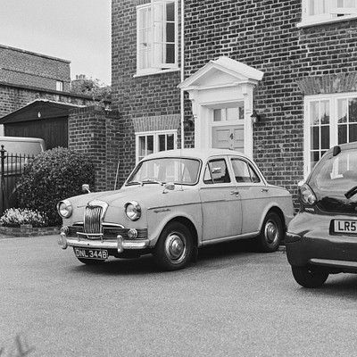 The image is a black and white photo of an old car parked in front of a house. The car appears to be a vintage model, possibly a Volkswagen Beetle. It is situated on the side of the road near a brick building with a white door.