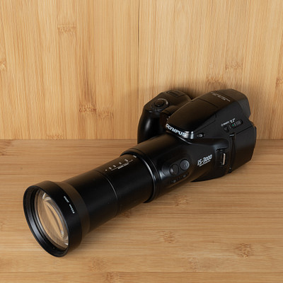 The image features a black camera with a lens on it, sitting on top of a wooden table. The camera is positioned in such a way that the lens is facing upwards, creating an interesting perspective. The wooden surface provides a natural and warm backdrop for this photography equipment.