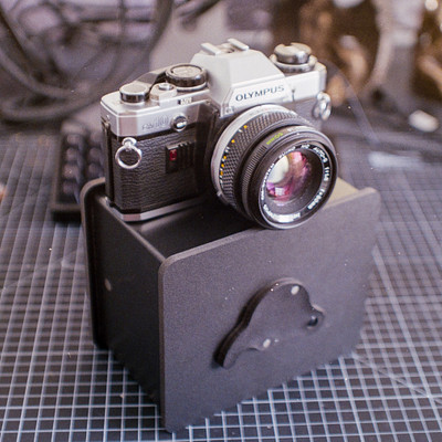 The image features a black box with a camera inside of it. The camera is an old-fashioned model, and it appears to be in good condition. There are several cords visible around the area where the camera is placed, possibly indicating that this setup is part of a photography or electronics workspace.