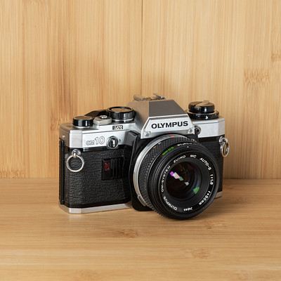 The image features an old-fashioned camera sitting on a wooden table. The camera is black and white, with a silver lens, giving it a vintage appearance. It appears to be an antique model, possibly from the 1950s or earlier.