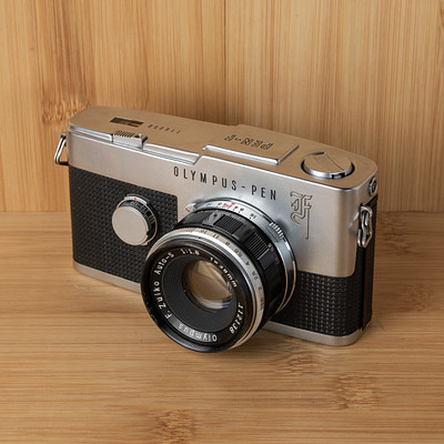 The image features a silver Olympus Pen camera sitting on top of a wooden table. The camera is placed in the center of the scene, drawing attention to its vintage design and classic appearance. The wooden surface beneath the camera adds a warm and natural touch to the overall composition.