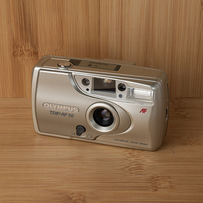 The image features a silver Olympus camera sitting on top of a wooden table. The camera is placed in the center of the scene, with its front facing towards the viewer. The wooden surface beneath the camera appears to be made from wood planks or slats, giving it a rustic and natural appearance.