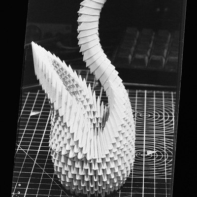The image features a unique, handmade sculpture of an animal, possibly a bird or a swan. It is made from paper and placed on top of a white plastic grid or a piece of cardboard. The artwork appears to be a three-dimensional representation of the animal, showcasing its intricate details and craftsmanship.